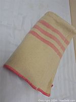 Folded beige wool blanket showing three pink horizontal stripes near one end and pink stitched edges.
