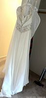 Full length front view of the white vintage wedding dress hanging on a hanger showing pleated flowing skirt and embellished shoulder straps.