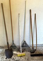 Photo of six garden tools leaning against a wall on concrete floor, showing shovel, two rakes, broom, hoe, and push broom.