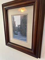 Framed art print of a narrow vintage street scene with people and storefronts, matted and set in a dark wood frame.