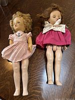 Two vintage dolls laying on a wood surface, one in light pink dress and bonnet, the other in dark pink dress with white collar. Both approximately 12 inches tall with eyes that open and shut.