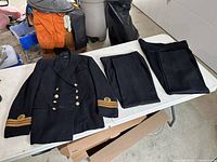 Full view of Royal Navy Reserve dress tunic with two pairs of matching black trousers laid next to it on table.