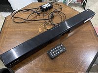 Samsung black sound bar laid on wooden surface with remote control and power cable visible.