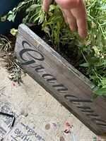 Side view of wooden planter box with 'Grandma' script and plants above showing leaves and soil.
