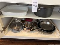Photo of cookware including saucepans, frying pans, ceramic baking dish, and large dark pot inside cabinet.
