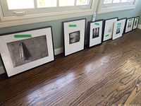 Framed black and white and color photographic prints lined up against wall on hardwood floor with window light