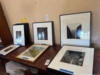 Five framed artworks displayed on a wooden tabletop against a neutral wall, showing a stack of black-framed photographic prints on left and two larger framed pieces on right, including a color painting in gold-tone frame