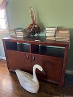 Front and left side view of wooden cabinet with books, feather arrangement on top, and wooden swan figurine on floor