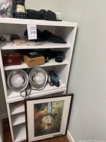 Front view of white shelving with assorted camera items and framed artwork leaning at base