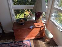 Wide view of the wooden chest with ceramic lamp and wooden duck sculpture beside it near window.