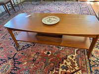 Full view of wooden coffee table on patterned rug with a decorative plate on top and wooden tray on the lower shelf