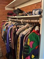 View of closet clothing hanging on rod; hats and folded fabric on upper shelf; wood paneling.