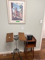 Full view of the dictionary stand and wooden side table with binocular case on top, with framed artwork on wall