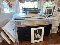 White work table holding a Masterpiece dry mounting press, sliding mat cutter and art prints on top. Black flat file cabinet with drawers underneath.