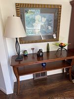 Photo of wooden entry table against wall with table lamp and various decorative items on top. Large framed floral wall art hangs above the table.