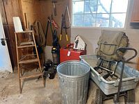 Wide shot showing the metal trash can, wooden step ladder, garden hoses on pegboard, garden tools hanging, vacuum cleaner, red toolbox, birdhouse, and lawn cart with folding chair seat.