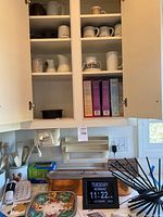 Overall kitchen counter and upper cabinet showing mugs, salt boxes, utensils, copper pan, decorative plates, digital clock, and plastic wrap dispenser.