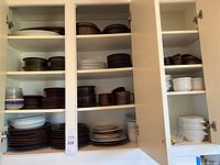 Three open kitchen cabinet doors showing multiple shelves filled with brown speckled stoneware and white ceramic plates, bowls, mugs, ramekins