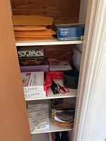 Overall view of closet shelf showing file folders, paper reams, boxes, stapler, scissors, and tape dispenser.