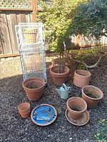 Photo shows arrangement of terracotta pots of various sizes on the ground with white metal wire rack behind holding more pots. Small watering can and tray with gardening tools are visible.