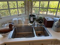 Kitchen sink area with three white ceramic canisters, a white ceramic pitcher, potted plant with bare branches, cactus, wooden bird decoy, wooden egg case, blue and white crock, and small wooden box around the sink.