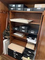Wood armoire interior with wooden shelves holding multiple black spiral-bound boxes and stacks of loose prints