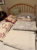 Bed with multiple quilts, blankets, and pillows piled on mattress including floral and solid cream quilts and pillows with fringe