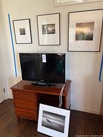 Samsung TV on wooden TV stand with three framed art prints on the wall and a framed print leaning against stand.