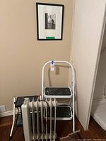 Photo of white two-step metal stool, black folding stool, oil heater, and framed photograph on wall