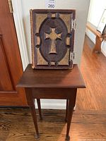 Bible and small wooden side table together, showing the Bible's front cover with central cross design and ornate detailing on the table top