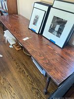 Long wooden table with four legs showing the top and under its surface where various items are stored, including framed photos and a TV.