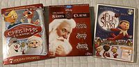 Front view of the three DVD cases included: The Original Christmas Classics tin, The Santa Clause DVD with Tim Allen, and The Elf on the Shelf Story DVD case.