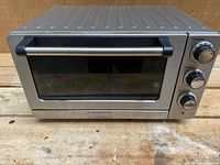 Front view of brushed stainless steel toaster oven with closed glass door, three control knobs visible on right side.