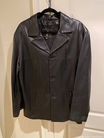 Front view of men's black leather coat hanging on a black hanger showing three-button closure and side pockets.