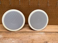 Pair of 9-inch round in-ceiling speakers with white frames and mesh covers, placed side by side on a wooden surface against a pegboard background.