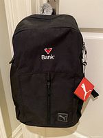 Front view of the black Puma backpack with the Bank logo embroidered on the front pocket and a Puma tag attached to the zipper.