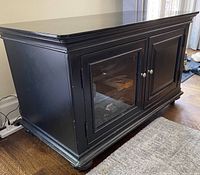 Front angle view of black wooden TV console showing one glass door and one solid door with silver knobs. Top shows some wear and paint chips on edges.