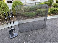 Front view of black curved mesh fireplace screen with brass handles next to tool stand with poker, brush, and shovel outdoors on gravel