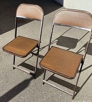 Two vintage folding chairs showing brown vinyl seats and backs and dark brown metal frames under outdoor lighting with shadows.