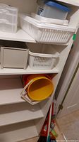 Shelves with white and beige plastic storage baskets, yellow plastic pail, and brooms leaning in the corner