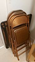 Four bronze metal folding chairs stacked together showing brown padded seats and backrests, with the folded black card table behind them.