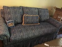 Full view of the paisley printed sofa showing rolled arms, skirted base, three back pillows, and one decorative fringed throw pillow.