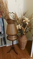 Full view of brown metal pitcher on wood tripod table and large faux floral arrangement in wicker-look vase beside it on floor