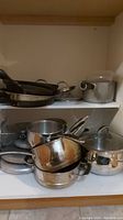 Two shelves inside a cupboard containing stacked pots and pans mostly in stainless steel and some non-stick, with several lids visible.