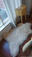 White sheepskin area rug laid on dark wood floor next to window and curtain, showing natural fur texture and shape.