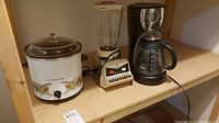 Photo showing Cornwall slow cooker, Osterizer blender, and Sunbeam coffee maker placed on a shelf.