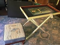 Foot stool next to hand painted serving tray on white folding stand on carpeted floor.