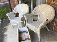 Two matching white wicker patio chairs on a concrete porch next to a sliding glass door with a small white wooden garden cart in front containing dried leaves.