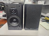 Two BIC Venturi bookshelf speakers, one with grille cover attached, black wood grain finish, sitting on white surface.
