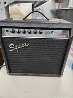 Front view of Squier Champ 15G guitar amplifier showing Squier logo, control knobs for gain, volume, treble, mid, bass, and input jack
